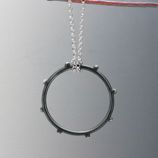 Firmament - Aloe - Oxidised Circle with Dots Silver Pendant. This exquisite piece boasts a 24mm diameter, or roughly 1 15/16 inches, and a thickness of 3mm.&nbsp;  Suspended from a 1.4mm silver Rolo chain with a secure spring ring clasp. Crafted from .925 sterling silver, it features an oxidized and high polished finish, adding depth and dimension to its intricate design. Choose your preferred chain length below