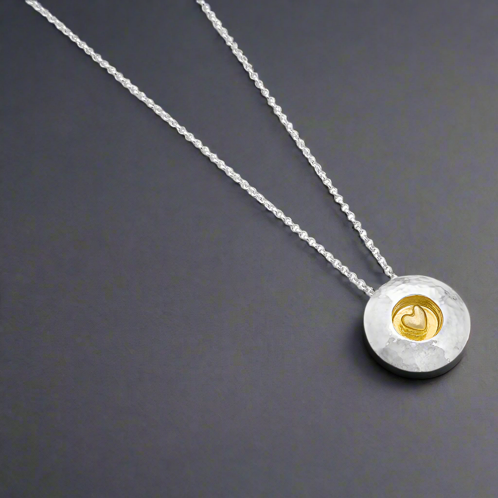 This pendant is a heartfelt design by Garrett Mallon, blending textured sterling silver with a sheltered 18ct gold vermeil heart. The domed silver disc symbolizes strength, while the golden heart represents love and connection, making it a meaningful choice for yourself or as a thoughtful gift. Croí Kinsale Jewellery Ireland.
