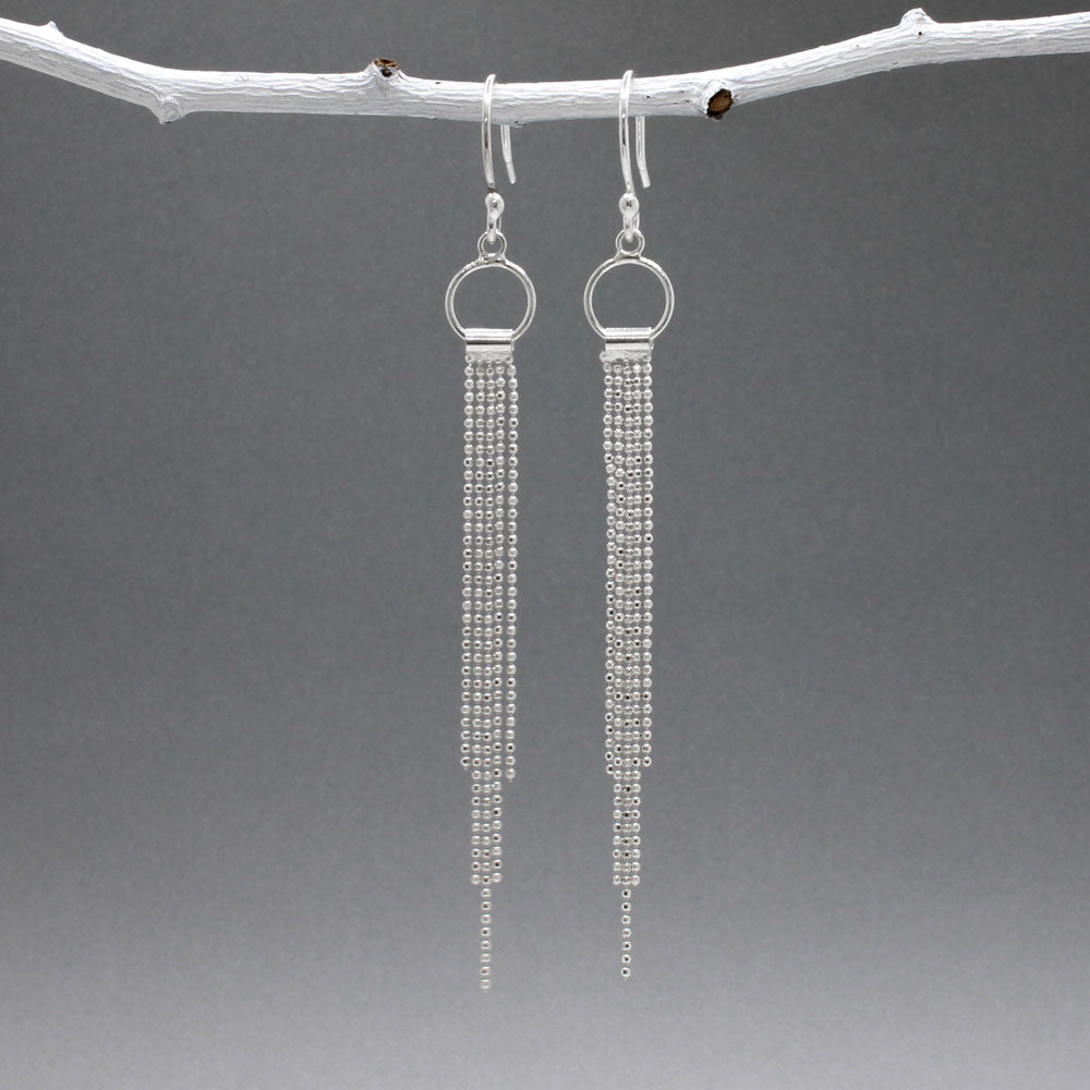 Áine Chains on Ring Silver Dangle Earrings. These handmade sterling silver earrings feature a delicate ring adorned with cascading chains, offering a unique and elegant look.  Features:  Material: Handmade sterling silver Design: Chains on a 9mm diameter ring Chain Width: 4mm at the widest area Length: 87mm from the top of the French wire hook Finish: Highly polished Closure: French wire hook for a secure and comfortable fit