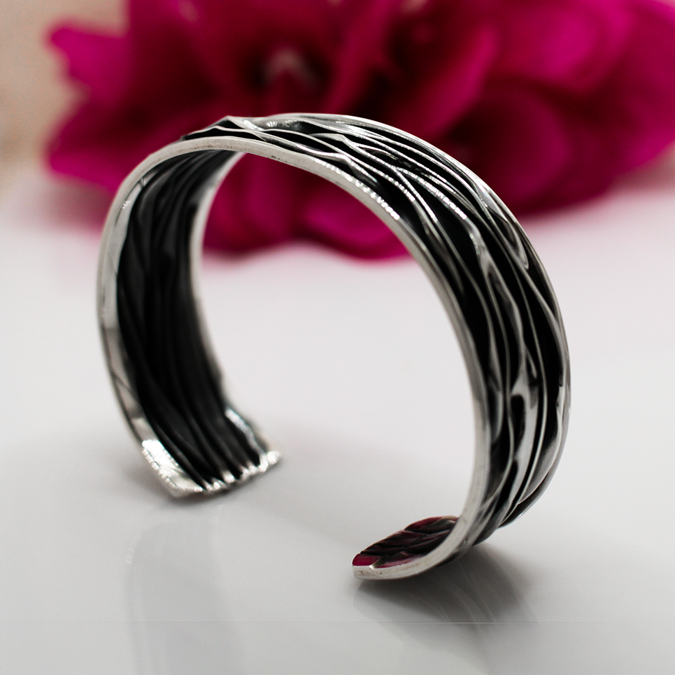 Uisce - Corrugated Oxidised Wide Cuff Silver Bracelet is meticulously handcrafted with sterling silver, boasting a striking oxidised finish. Each cuff features a unique wave pattern, enhanced by polished silver and oxidised details, ensuring that no two pieces are exactly alike.