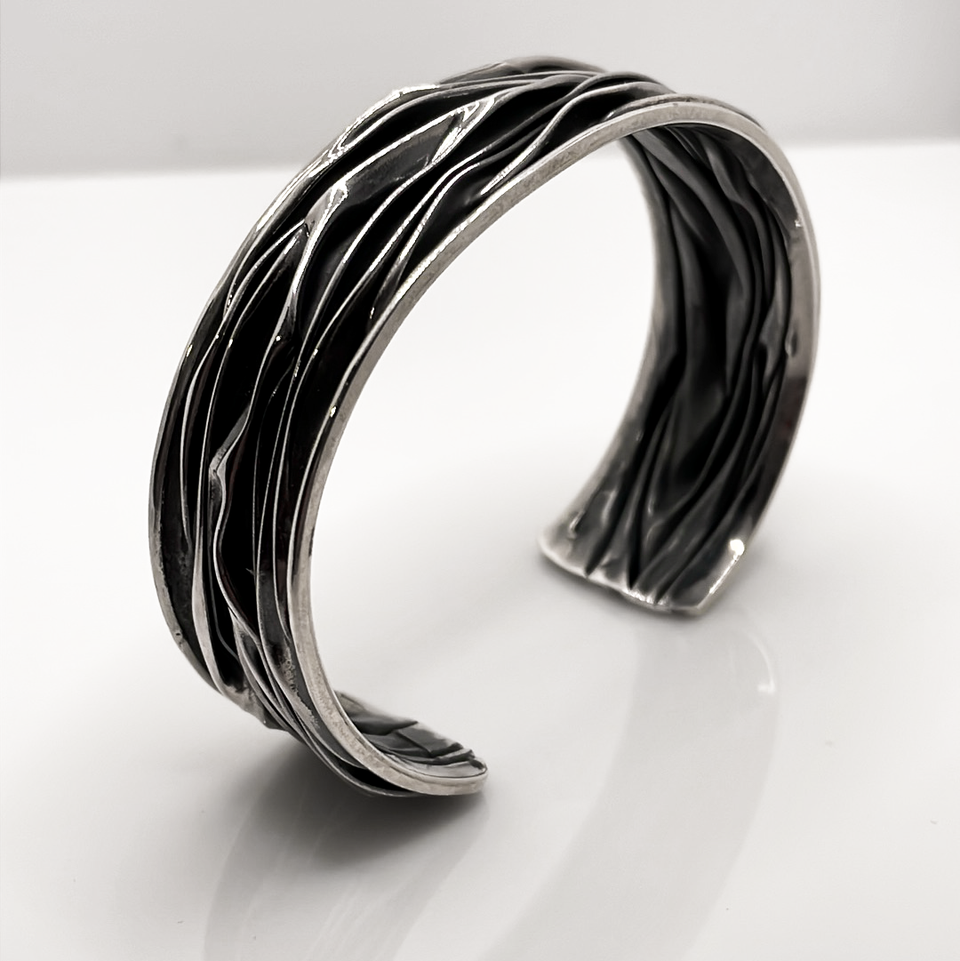 Uisce - Corrugated Oxidised Wide Cuff Silver Bracelet is meticulously handcrafted with sterling silver, boasting a striking oxidised finish. Each cuff features a unique wave pattern, enhanced by polished silver and oxidised details, ensuring that no two pieces are exactly alike. Ireland