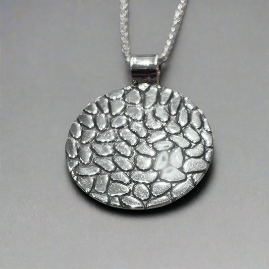 The Camila Disc Oxidised Silver Pendant features a bold, circular design with a striking cobblestone pattern. Crafted from .950 sterling silver, this pendant blends an oxidized texture with a highly polished silver finish.

Material: .950 sterling silver with an oxidized cobblestone pattern and highly polished finish
Dimensions:
Diameter: 30mm (1 3/16 inches)
Chain: Includes a 2.5mm sterling silver Rolo chain with a lobster claw clasp (select your preferred length)
Bail: Tube bail fits up to a 5mm chain (3/