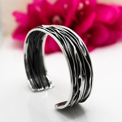 Uisce - Corrugated Oxidised Wide Cuff Silver Bracelet is meticulously handcrafted with sterling silver, boasting a striking oxidised finish. Each cuff features a unique wave pattern, enhanced by polished silver and oxidised details, ensuring that no two pieces are exactly alike.