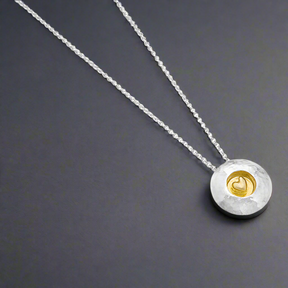 This pendant is a heartfelt design by Garrett Mallon, blending textured sterling silver with a sheltered 18ct gold vermeil heart. The domed silver disc symbolizes strength, while the golden heart represents love and connection, making it a meaningful choice for yourself or as a thoughtful gift. Croí Kinsale Jewellery Ireland.