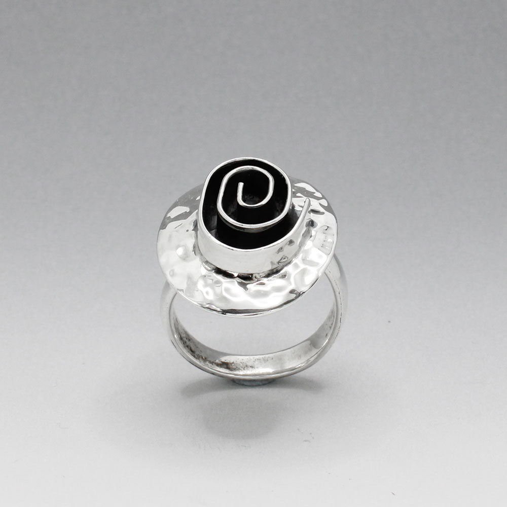 Tara - Oxidised Spiral On Hammered Oval Silver Ring  Discover the unique charm of our Tara ring, featuring an exquisite oxidised spiral design set on a hammered oval silver base. This striking piece measures 20mm wide by 25mm long, with the spiral itself measuring 12mm wide by 17mm long.  The adjustable band ensures a perfect fit, ranging from size 7 (European size 54) to size 9 (European size 59). Crafted from solid sterling silver, the 4mm wide band offers durability.