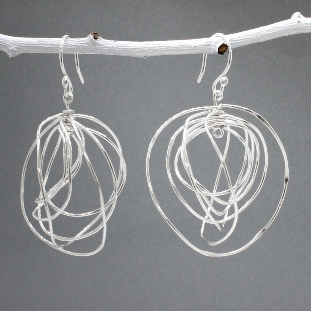 Tara - Twisting Multi-Circle Silver Earrings - Dangle  Design: Seven graduated hoops interconnected, creating an organic circle shape  Movement: Rotates when worn, adding whimsical and dynamic movement Features: Lightweight, highly polished silver Fastening: Sterling silver French wire Measurements: Width: 37 MM at its widest point Length: 55 MM from the top of the French wire. Ireland.