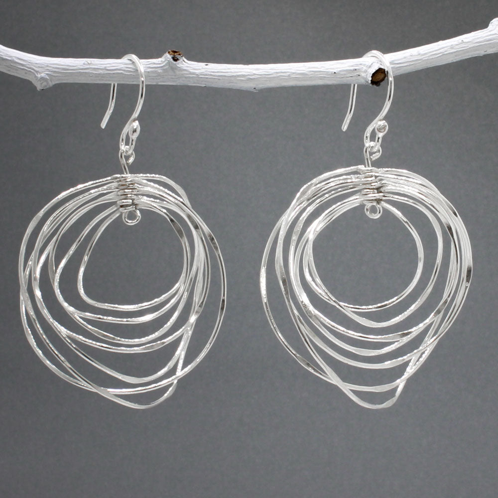 Tara - Twisting Multi-Circle Silver Earrings - Dangle  Design: Seven graduated hoops interconnected, creating an organic circle shape  Movement: Rotates when worn, adding whimsical and dynamic movement Features: Lightweight, highly polished silver Fastening: Sterling silver French wire Measurements: Width: 37 MM at its widest point Length: 55 MM from the top of the French wire. Kinsale