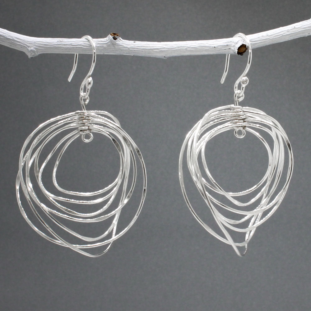 Tara - Twisting Multi-Circle Silver Earrings - Dangle  Design: Seven graduated hoops interconnected, creating an organic circle shape  Movement: Rotates when worn, adding whimsical and dynamic movement Features: Lightweight, highly polished silver Fastening: Sterling silver French wire Measurements: Width: 37 MM at its widest point Length: 55 MM from the top of the French wire. Kinsale jewellery store.